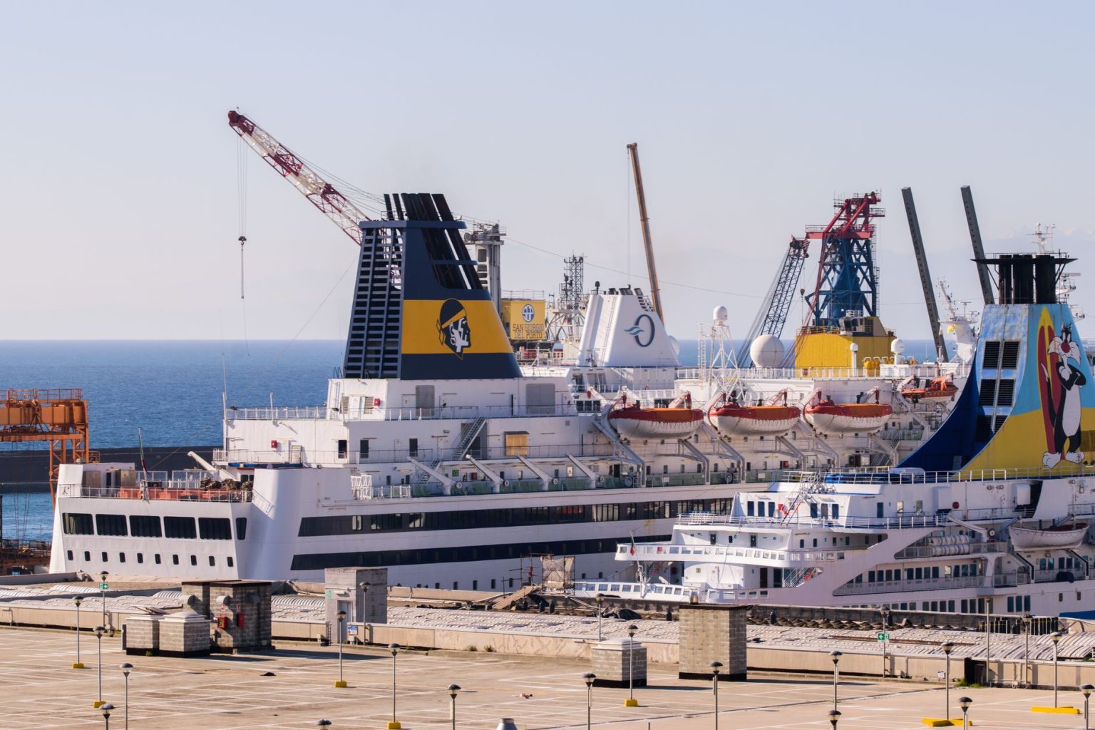 Navios de cruzeiro atracados em um porto, com guindastes ao fundo e céu claro.