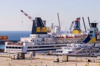 Navios de cruzeiro atracados em um porto, com guindastes ao fundo e céu claro.