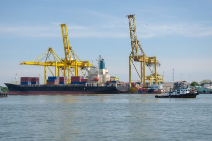 Navio cargueiro atracado em um porto industrial, cercado por guindastes e contêineres.