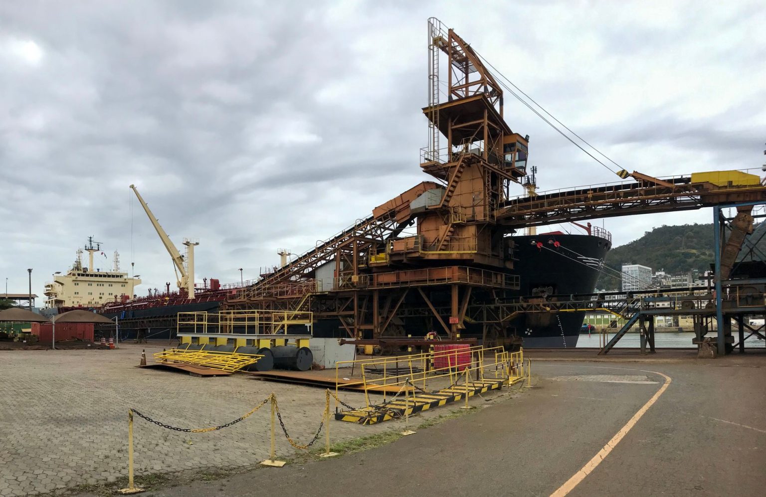Navios e guindastes operam em um porto industrial junto a uma estrutura de carregamento.