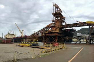 Navios e guindastes operam em um porto industrial junto a uma estrutura de carregamento.