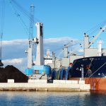Navio cargueiro com guindastes em um porto industrial durante o carregamento de minerais.