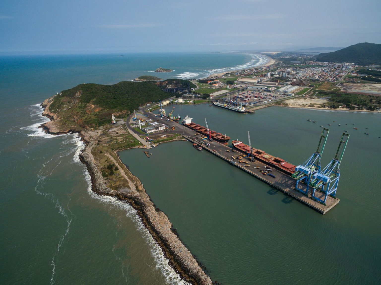 Vista aérea do Porto de Imbituba, com cais, guindastes, embarcações e a costa brasileira