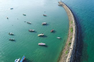 Vista aérea de um longo quebra-mar em construção no mar, com embarcações ao redor.