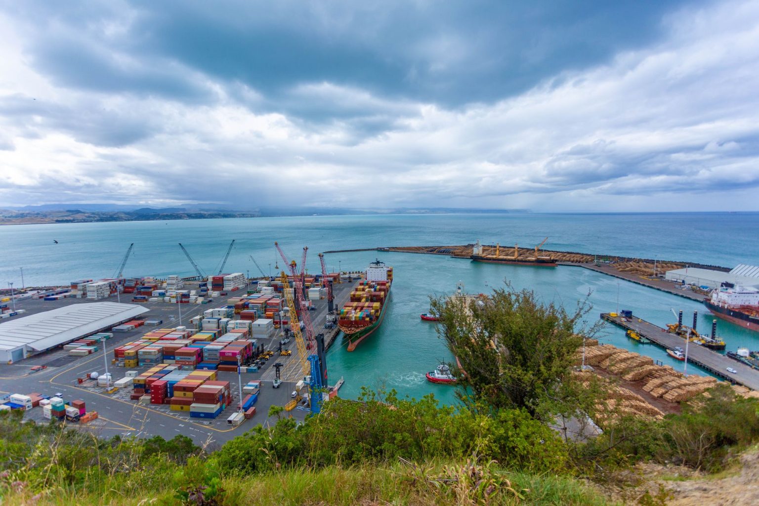 Vista de um porto marítimo movimentado, com navios cargueiros, contêineres e guindastes sob um céu nublado.