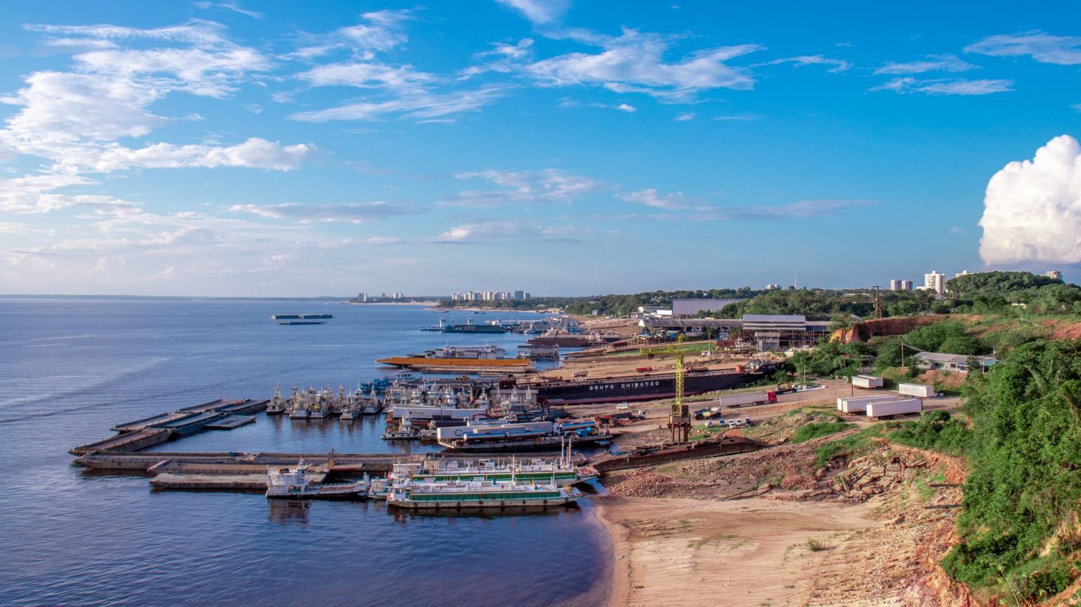 Vista aérea de um porto às margens do Rio Negro, em Manaus, com embarcações atracadas e a cidade ao fundo.