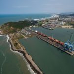 Vista aérea de um porto brasileiro, com cais, guindastes, embarcações e trecho do litoral.