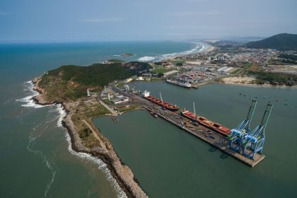 Vista aérea de um porto brasileiro, com cais, guindastes, embarcações e trecho do litoral.
