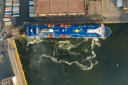 Vista aérea de um navio de carga atracado em um porto industrial, com estruturas do cais visíveis.