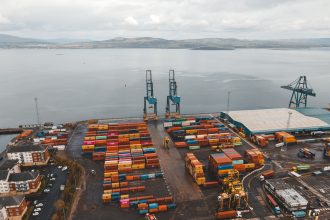 Vista aérea de um terminal portuário movimentado, com guindastes e contêineres coloridos.