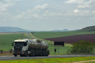 Caminhão de carga trafega por uma rodovia entre extensos campos agrícolas.